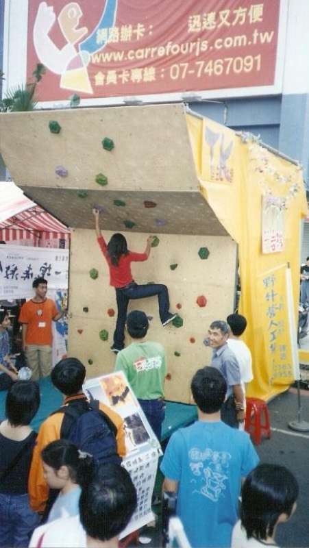 戶外廣場舉辦的小型攀岩體驗活動，一名女子正在攀岩，眾人在旁圍觀