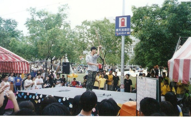 TK在校園戶外舞台上持長棍表演，台下聚集眾多學生觀看
