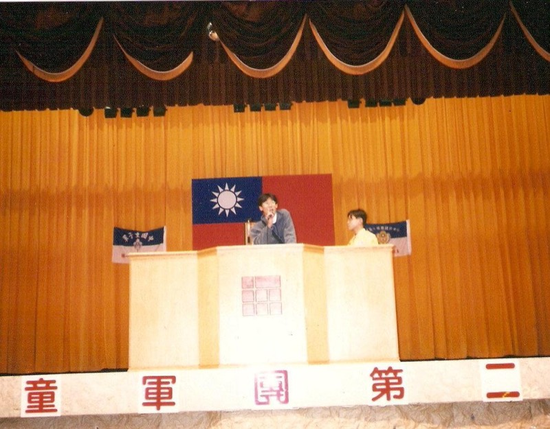 年輕TK在禮堂講台上持麥克風發言，背景懸掛中華民國國旗與童軍旗幟。